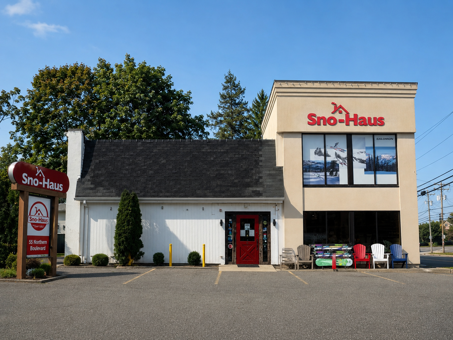 Sno-Haus Greenvale ski store building exterior with a beige and white facade, red doors and signage, a parking lot, trees, and a blue sky.