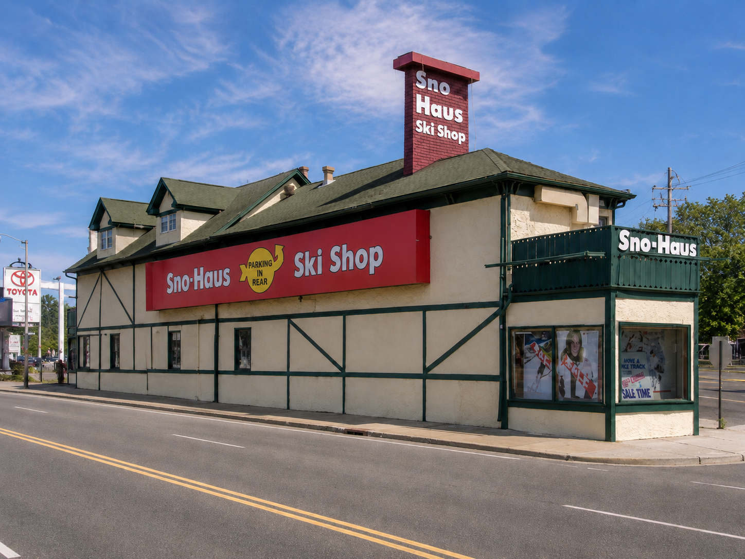 Sno-Haus Ski Shop Hempstead building with cream-colored timber framing, dark green trim, and a prominent red sign on the corner. The building is situated on a street corner with a paved road and double yellow lines in the foreground, under a blue sky with light clouds.