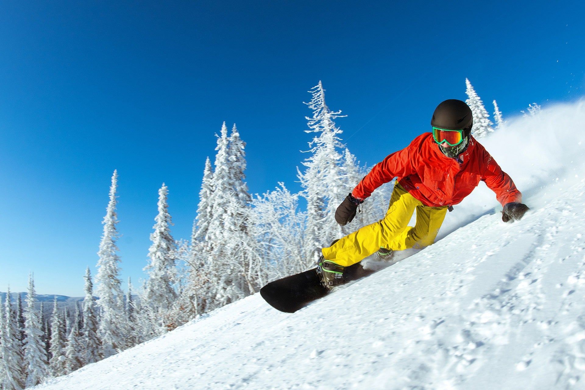 Snowboarder carving on snowy mountain wearing red jacket and yellow pants.