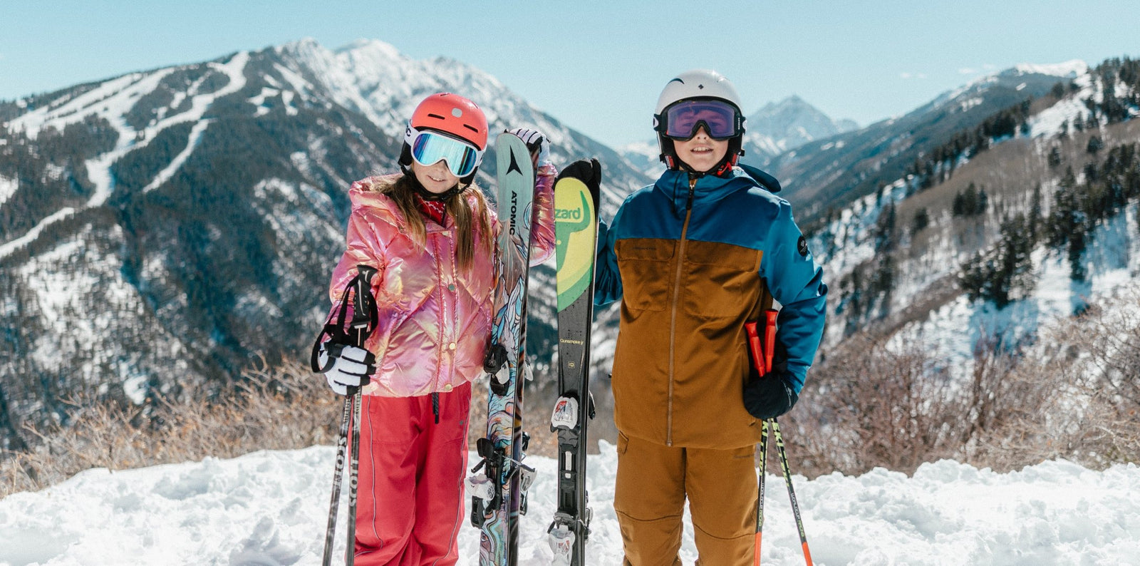 Two young skiers in helmets and goggles holding skis and poles on a snowy mountain