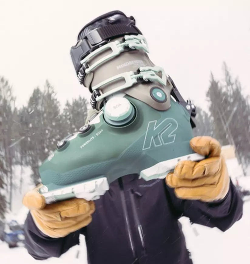 Person holding a green ski boot with 'K2' branding in a snowy landscape