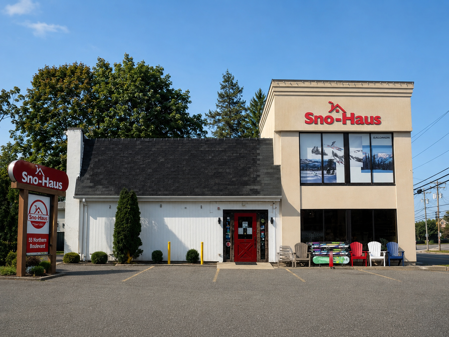 Sno-Haus Greenvale ski store building exterior with a beige and white facade, red doors and signage, a parking lot, trees, and a blue sky.
