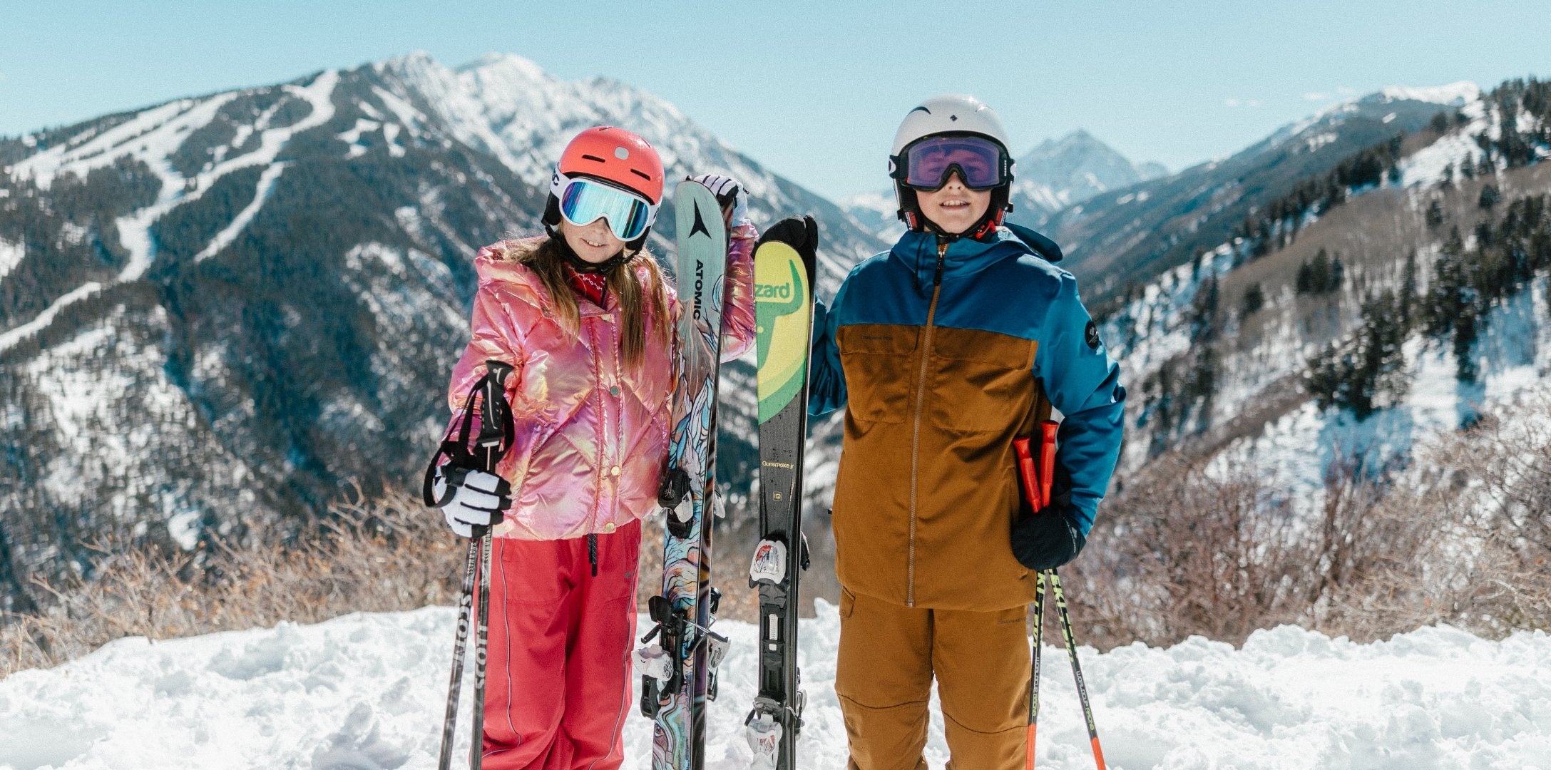 Two young skiers in helmets and goggles holding skis and poles on a snowy mountain
